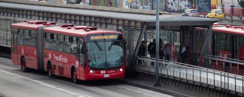 Red bus in font of covered pedestrian walkway/station. Partially visible bus on the other side of the station, a few pedestrians visible near the end of the covered walkway.