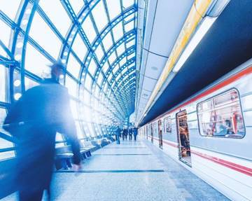 Walkway with vaulted transparent covering. People walk along it, and one side of a train or bus is visible to the right.